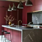 A stylish kitchen with sleek burgundy cabinets and a deep charcoal island topped with marble featuring prominent white veining. The kitchen’s sophisticated palette is complemented by matte black pendant lights hanging above the island, with their industrial style adding a modern touch. A brass faucet stands out on the island, paired with black and gold accents for an elegant look. Minimalist black bar stools with cushioned seats provide seating, while a gold vase with delicate branches adds an organic touch to the refined, contemporary space.