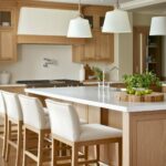 Charming kitchen with light oak cabinetry and integrated lighting, featuring a white quartz island with stylish white upholstered bar stools. Includes elegant pendant lighting, modern kitchen appliances, and a minimalist decor with natural green accents, creating a warm and welcoming atmosphere.