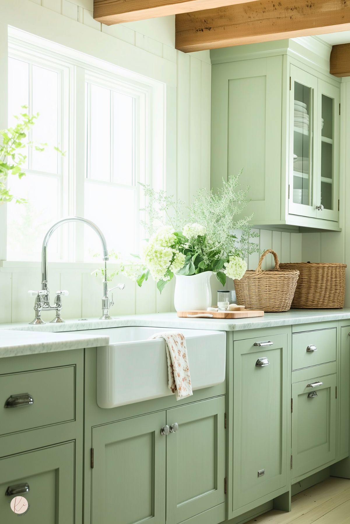 A bright and airy kitchen with soft sage green cabinetry and a white farmhouse sink. The countertop features a white pitcher filled with fresh hydrangeas and greenery, along with woven baskets adding natural texture. Sunlight streams through large windows, highlighting the polished chrome faucet and beadboard walls, creating a charming and tranquil cottage-inspired space.