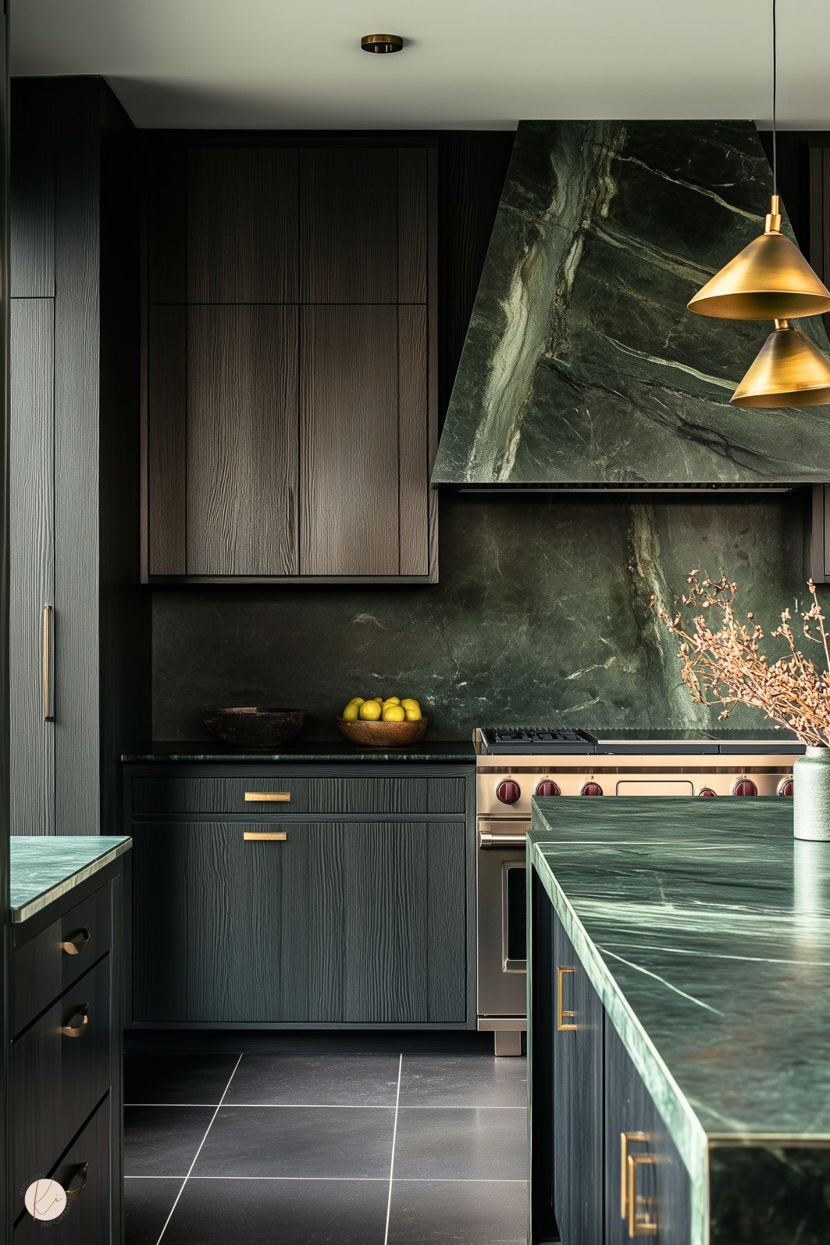 A luxurious kitchen with dark wood cabinetry and a striking green marble backsplash and countertop that exude sophistication. Brass hardware and pendant lights add a touch of warmth and elegance, contrasting beautifully with the deep, moody tones. The seamless marble range hood serves as a bold centerpiece, while minimalistic decor, including a bowl of lemons and dried branches, maintains a clean and refined aesthetic.