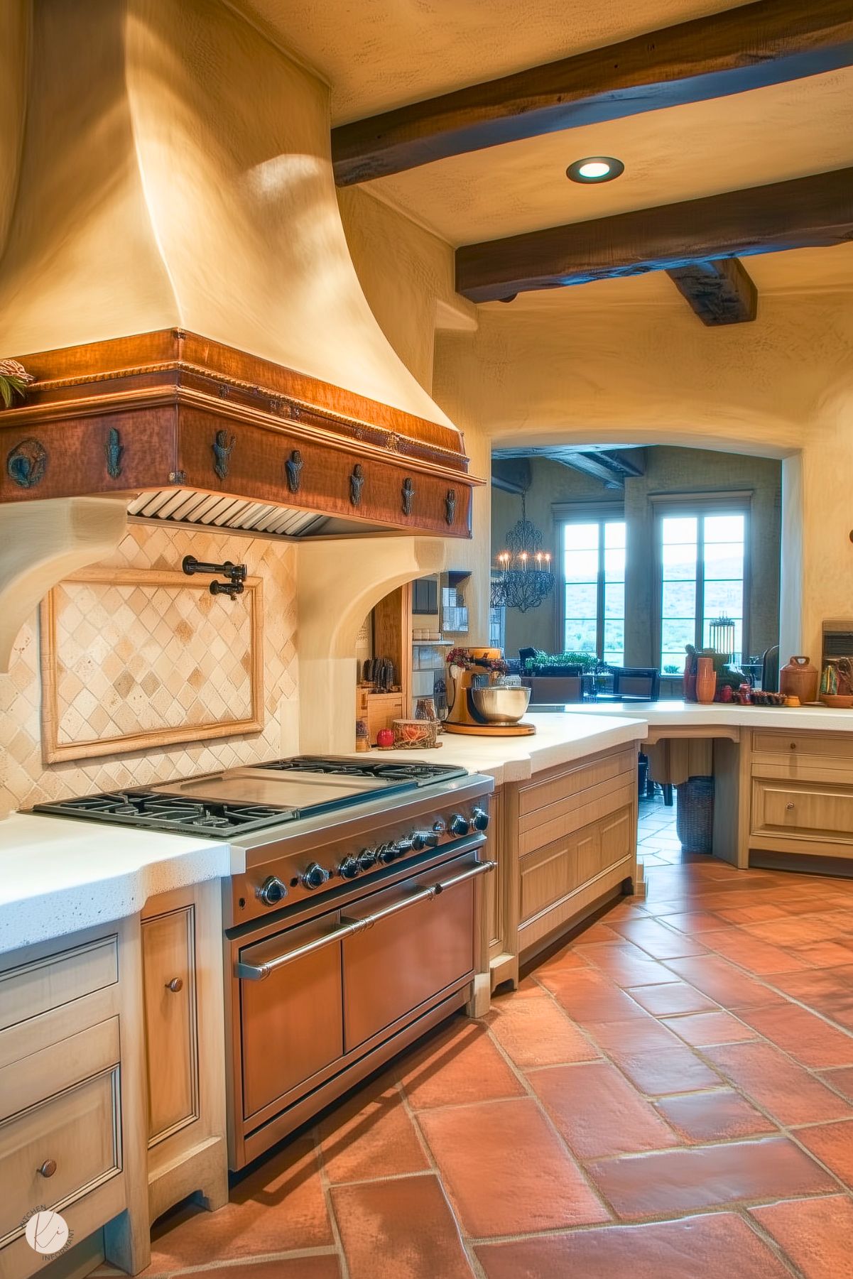 A Tuscan-inspired kitchen featuring a stunning copper range hood with decorative accents, set above a professional-grade oven. The backsplash is composed of warm, textured beige tiles framed by a simple yet elegant design. Custom wood cabinetry with intricate detailing complements the curved white countertops. Terracotta tile flooring enhances the rustic charm, while exposed wooden beams and arched doorways add depth and character to the inviting space. Large windows in the background bring in natural light, completing the Mediterranean ambiance.