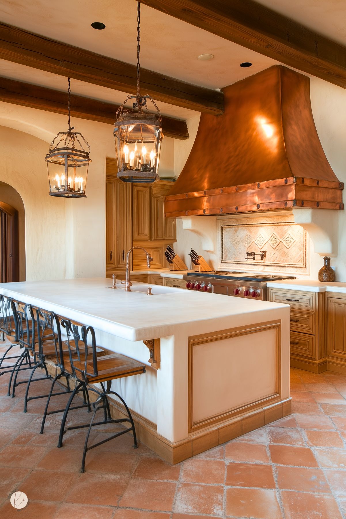 A stunning rustic kitchen featuring a large white island with intricate wood detailing and a polished copper sink. Above the island hang two lantern-style chandeliers, providing warm lighting and a touch of elegance. The centerpiece is a striking copper range hood with rivet details, perfectly complementing the wood cabinetry and tiled backsplash. Terracotta floor tiles enhance the earthy tones, while wrought iron barstools with wooden seats add a farmhouse-inspired charm. The space is both luxurious and inviting.