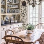 Rustic kitchen with a vintage wooden dining table, spindle-back chairs, a botanical wallpaper accent, and open shelving displaying blue and white china. A chandelier adds an elegant touch, with natural light streaming in through large windows.