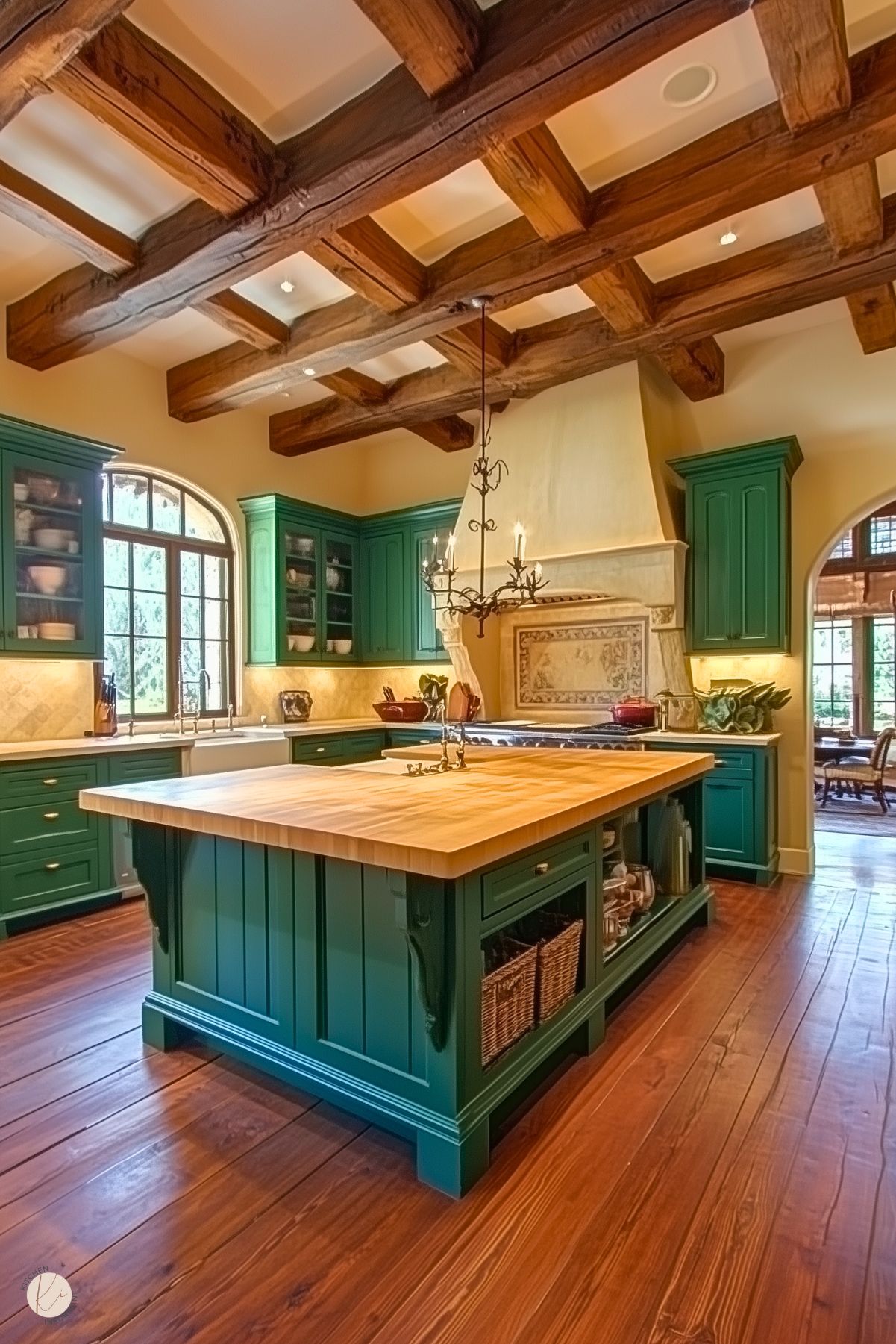 A traditional kitchen with rich green cabinetry, a large wood-topped island, exposed wooden ceiling beams, a farmhouse sink, and an ornate chandelier over the island.