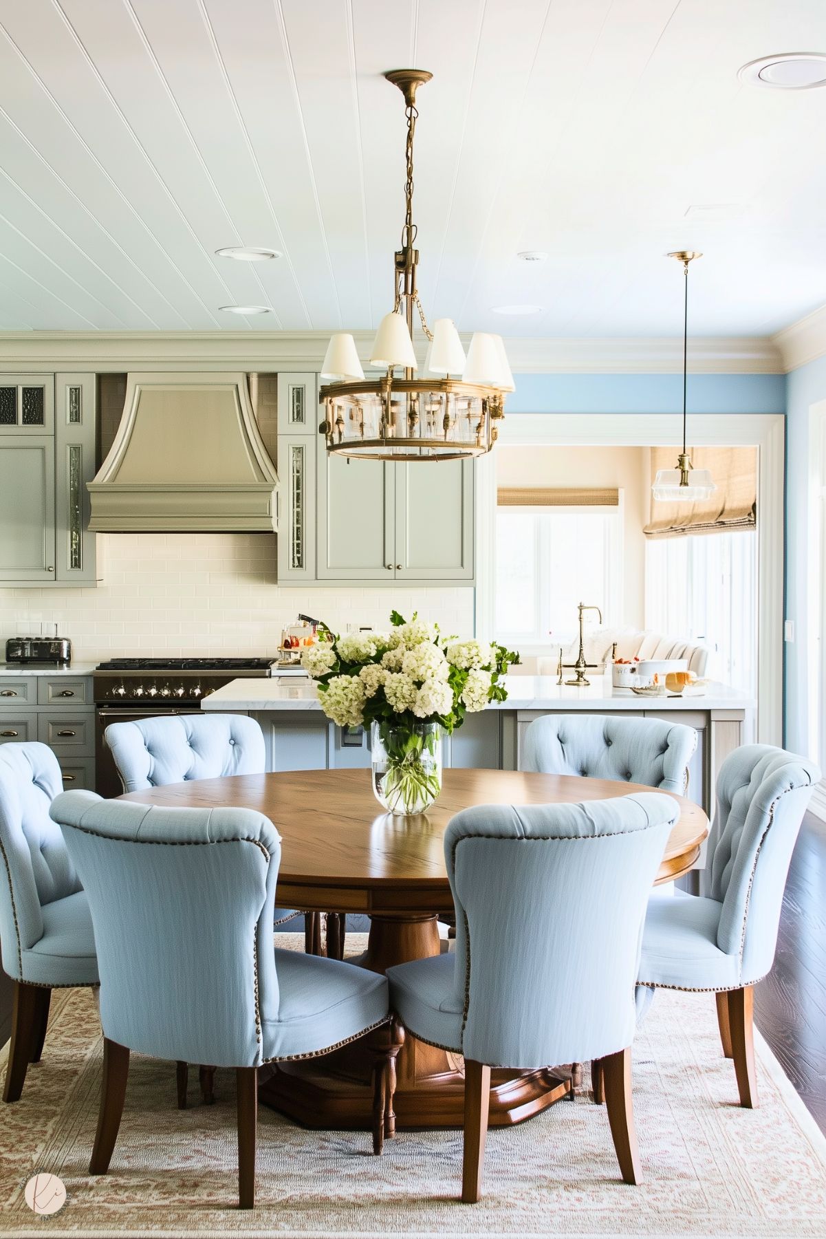 A cozy, elegant kitchen featuring soft blue cabinetry, a round wooden dining table, and plush blue upholstered chairs. A vase of fresh white flowers serves as the centerpiece, while a classic chandelier adds charm above. The bright, airy space is complemented by light walls, natural light, and subtle decorative accents.