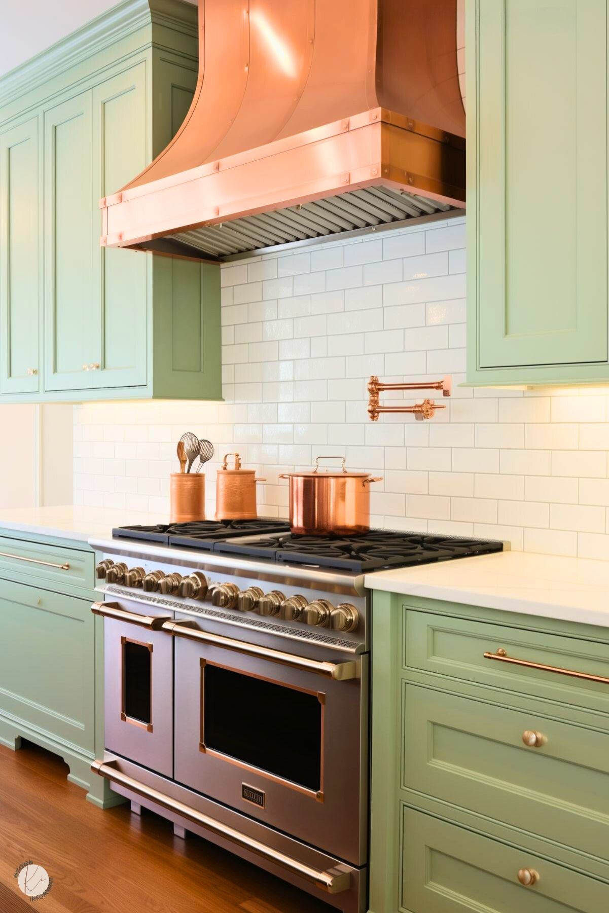 A sophisticated kitchen featuring sage green cabinetry with polished nickel hardware. A stunning copper range hood takes center stage above a stainless steel professional stove, accented by copper cookware. The backsplash is composed of classic white subway tiles, complemented by a copper pot filler. Bright under-cabinet lighting and a warm wood floor complete the elegant, timeless design.