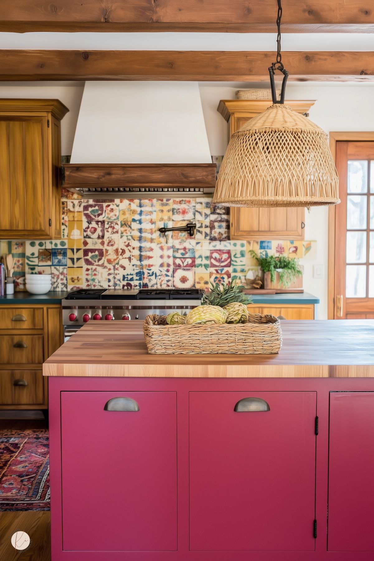 A rustic kitchen featuring a burgundy island with a butcher block countertop and black cup pull handles. Above the island, a woven rattan pendant light adds texture and warmth. The backsplash showcases colorful, patterned tiles, complementing the wooden cabinetry and exposed ceiling beams. A woven basket with fresh produce decorates the island, and a vintage-style range with red knobs adds a functional yet charming focal point.