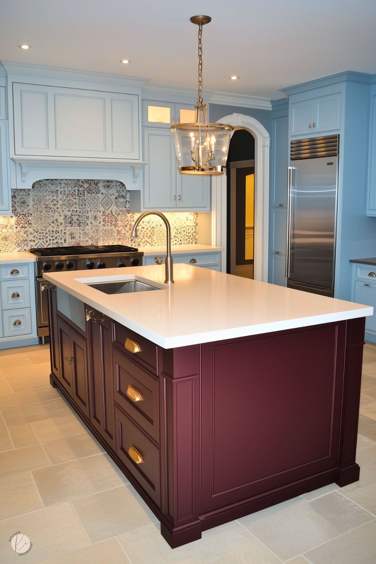 A bright kitchen featuring a deep burgundy island with paneled detailing, a white quartz countertop, and brass hardware. The surrounding cabinetry is painted in soft blue, complemented by a patterned tile backsplash that adds a touch of charm. A brass pendant light with glass accents hangs over the island, enhancing the warm, inviting atmosphere. Stainless steel appliances and cream floor tiles complete the elegant yet functional design.