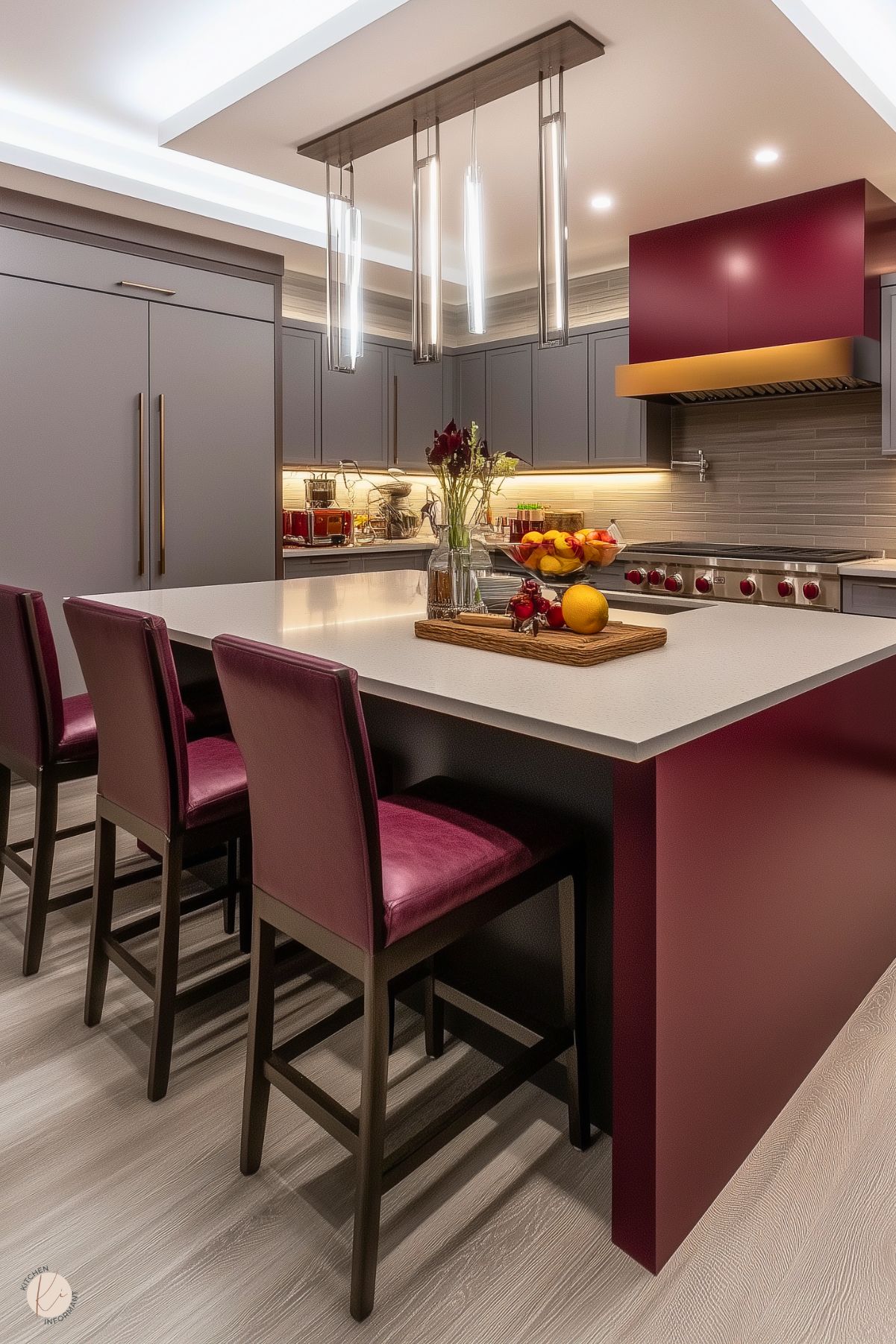 A contemporary kitchen with a burgundy island topped with a sleek white countertop. The island is surrounded by matching burgundy leather bar stools, creating a cohesive design. Modern pendant lights with glass elements hang above, adding elegance. The gray cabinetry, integrated appliances, and tiled backsplash contrast beautifully with the warm burgundy accents. A decorative range hood in burgundy and gold serves as a bold focal point.