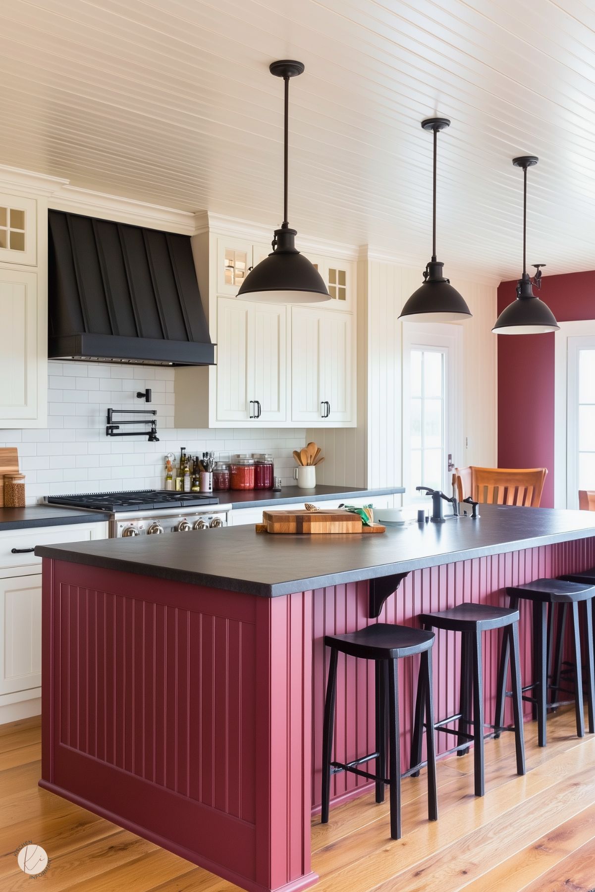 A charming farmhouse-style kitchen featuring a burgundy beadboard island with a black countertop and matching black bar stools. The space includes cream cabinetry, a white subway tile backsplash, and a striking black range hood. Three industrial-style black pendant lights hang above the island, adding contrast and character. Natural wood flooring and warm burgundy walls complete the cozy and inviting aesthetic.