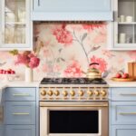 A kitchen featuring powder blue cabinets with brass hardware, a stainless steel and brass gas range with matching knobs, and a floral backsplash in shades of pink and gray. The countertop is white, styled with a vase of pink flowers, a gold kettle, and a wooden tray with red apples.