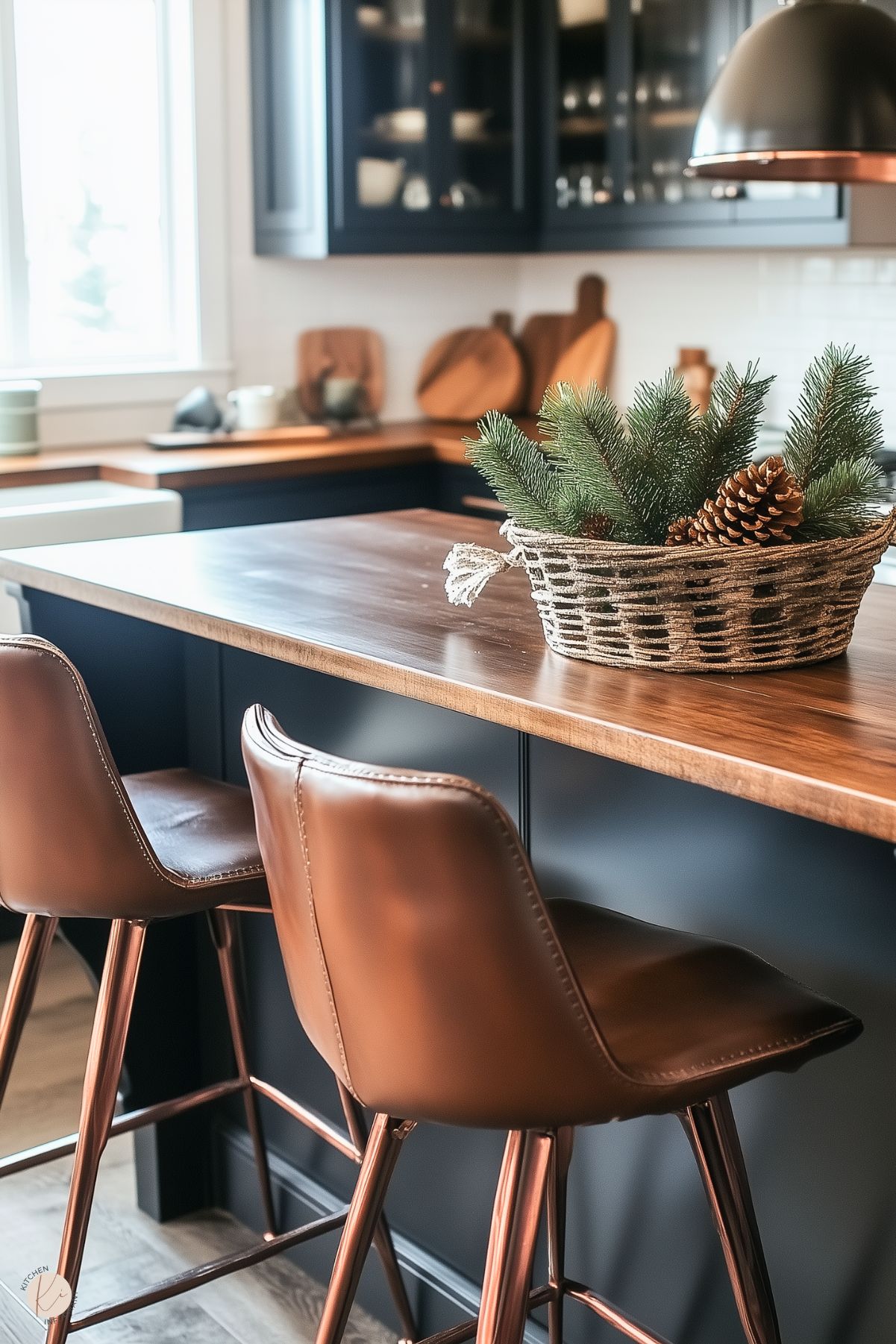 A cozy, modern kitchen featuring a wooden island countertop paired with dark cabinetry. Two leather barstools with copper accents sit at the island, adding warmth and elegance. A woven basket centerpiece with pine branches and pinecones brings a festive, natural touch. The background highlights glass-front cabinets, wooden cutting boards, and a soft white backsplash, creating a harmonious blend of rustic and contemporary styles.