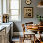 A charming, traditional kitchen nook with warm gray walls and white cabinetry. A small wooden dining table with matching chairs sits atop an ornate vintage rug. A bowl of fruit and a potted plant add natural touches to the space. The window features a simple beige Roman shade, while a copper pendant light hangs above. Decor includes a wall clock and framed artwork, enhancing the cozy, classic atmosphere.