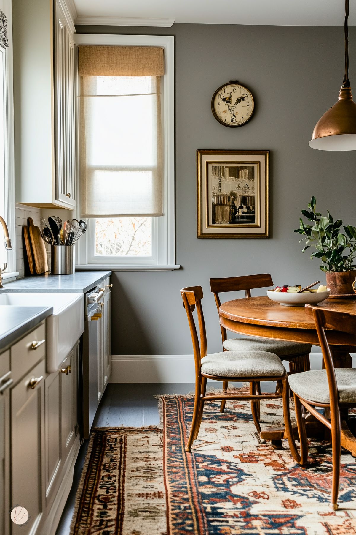 A charming, traditional kitchen nook with warm gray walls and white cabinetry. A small wooden dining table with matching chairs sits atop an ornate vintage rug. A bowl of fruit and a potted plant add natural touches to the space. The window features a simple beige Roman shade, while a copper pendant light hangs above. Decor includes a wall clock and framed artwork, enhancing the cozy, classic atmosphere.