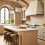 A charming kitchen with a vaulted ceiling featuring exposed wooden beams and a soft red brick pattern. The space includes a central light wood island with turned legs, a white countertop, and leather barstools with nailhead trim. Elegant lantern-style pendant lights hang above the island. The cabinetry is cream-colored with glass-fronted upper cabinets, and the custom range hood matches the soft tones. A large arched black-framed window brings in natural light, highlighting the bright and inviting design.