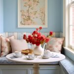 A charming kitchen nook with soft blue walls, a built-in bench featuring striped and floral cushions, and a round white pedestal table. A white pitcher filled with vibrant red flowers acts as a centerpiece, accompanied by teacups, saucers, and a plate of cookies. A framed floral artwork decorates the wall above, and natural light streams in through a nearby window.