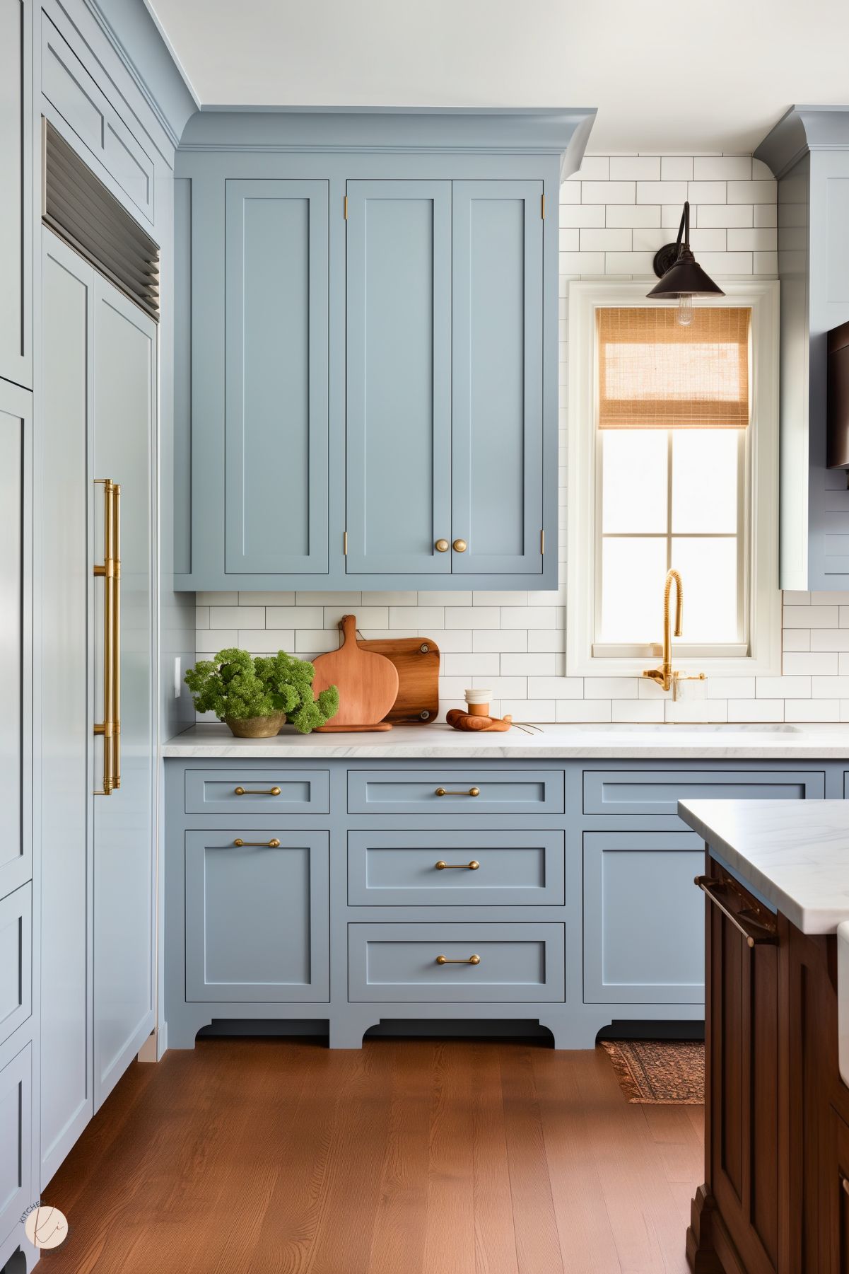 A farmhouse-style kitchen with soft blue shaker cabinets, brass hardware, and a white subway tile backsplash. A white marble countertop holds wooden cutting boards, a brass faucet, and a potted leafy green plant near a window with a woven shade. Warm wood floors and a rich-toned island contrast the light palette, creating a cozy yet elegant space.