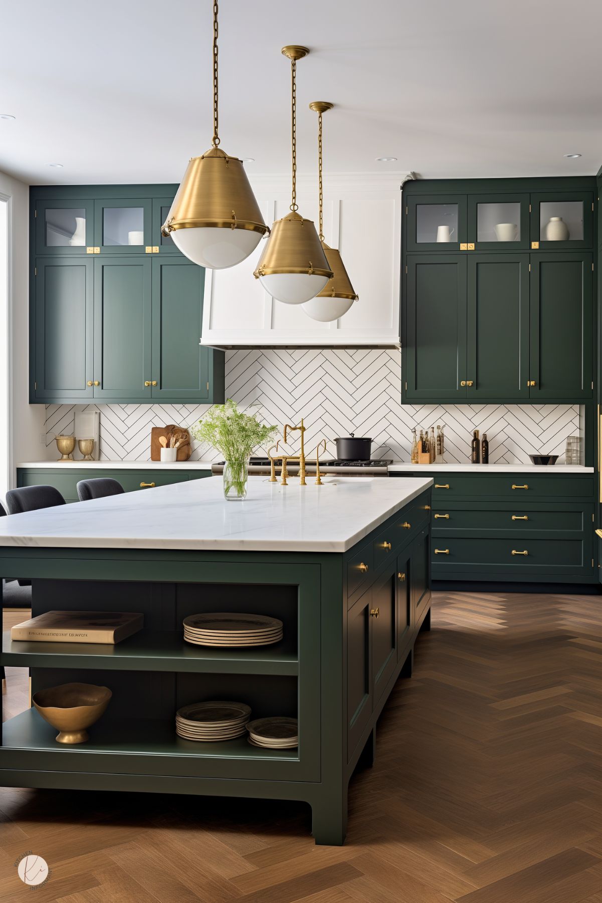 A modern kitchen featuring deep forest green cabinetry paired with brass hardware. A large central island with open shelving and a white countertop stands in the foreground. Above, three brass pendant lights hang over the island, adding warmth. The backsplash showcases a white herringbone tile pattern, while the flooring is a chevron wood design, tying the look together with elegance and texture.