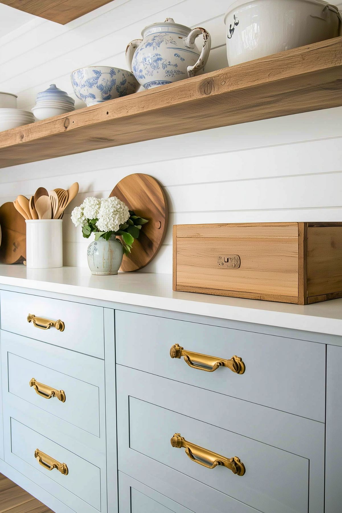 A charming French cottage kitchen featuring soft blue cabinetry with elegant brass handles. A white countertop is adorned with a rustic vase of white hydrangeas, wooden utensils in a ceramic holder, and a wooden storage box. Above, a natural wood floating shelf displays delicate blue-and-white ceramics and bowls, enhancing the rustic yet refined aesthetic. Shiplap walls add texture and warmth to the inviting space.