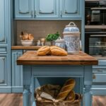 A charming French cottage kitchen with powder blue cabinets featuring gold accents and a warm wooden countertop. The centerpiece is a small island adorned with freshly baked bread and blue-and-white porcelain jars. A wicker basket filled with baguettes is tucked neatly underneath the island. The soft subway tile backsplash and cozy lighting add to the inviting ambiance of the space.