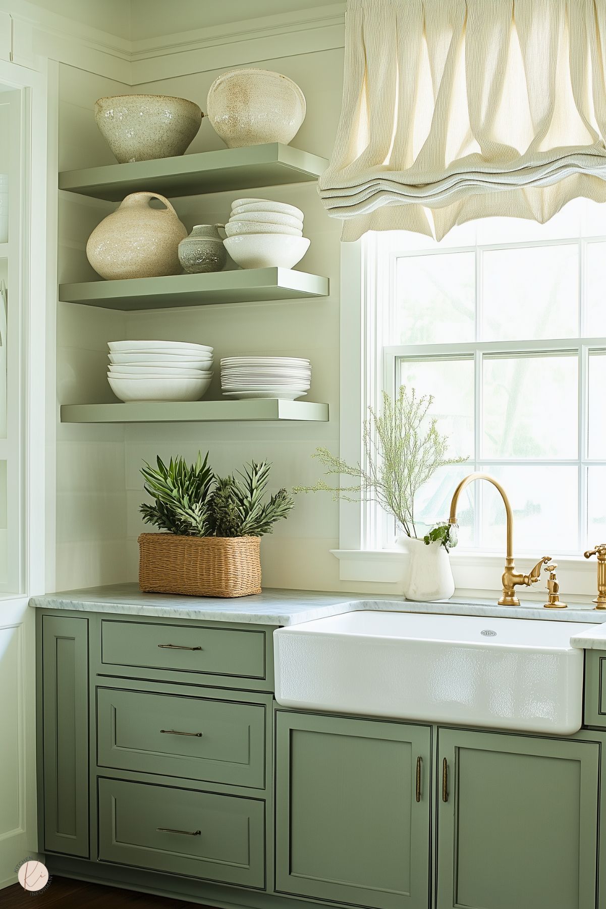 A serene French cottage kitchen with sage green cabinetry and a marble countertop. A farmhouse sink with a polished brass faucet is framed by a large window, adorned with soft, gathered fabric shades. Floating shelves display minimalist pottery and stacked dishes, while a woven basket filled with fresh greenery adds texture and charm. The overall design balances rustic and elegant elements for a warm, inviting space.