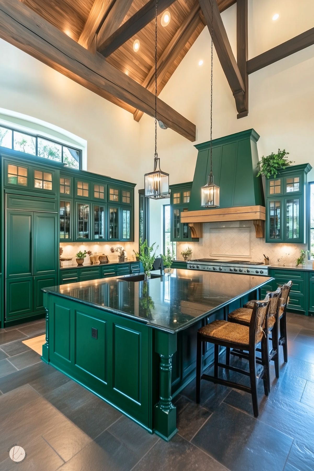 A spacious kitchen with rich green cabinetry and a large island featuring a polished black countertop. The vaulted ceiling with exposed wooden beams enhances the room's grandeur, while pendant lanterns provide warm lighting. Glass-front cabinets display elegant dishware, and a wooden range hood adds warmth and texture. Stone flooring and natural greenery accents complete the luxurious, earthy ambiance.