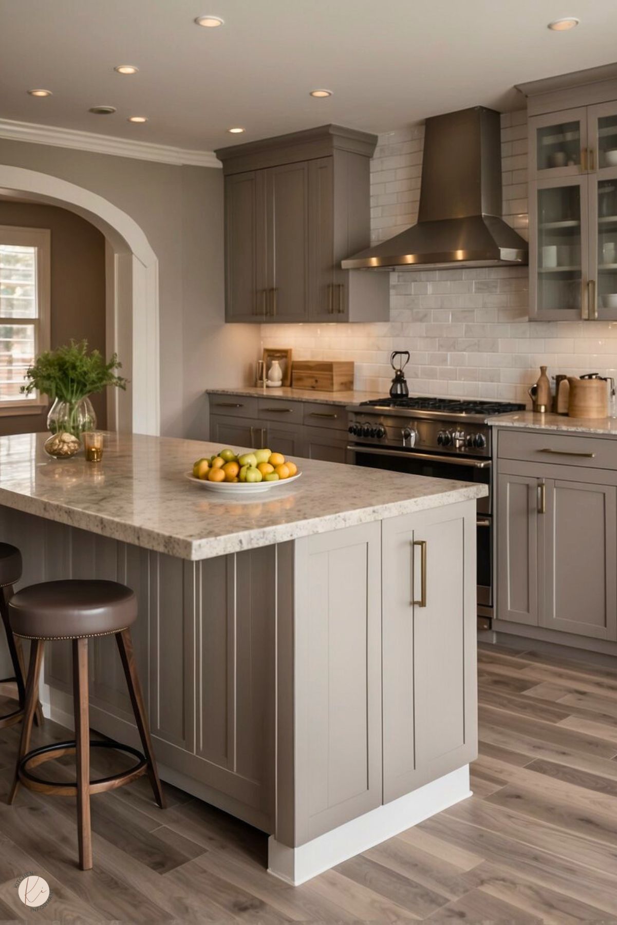 The image showcases a warm and inviting greige kitchen with a large island featuring a speckled stone countertop and shaker-style cabinetry. The island includes bronze hardware and is paired with two leather-upholstered bar stools. The background displays matching greige cabinetry, a stainless steel range hood, and a white subway tile backsplash. The room is illuminated by recessed lighting, and the hardwood flooring adds a cozy, natural touch.