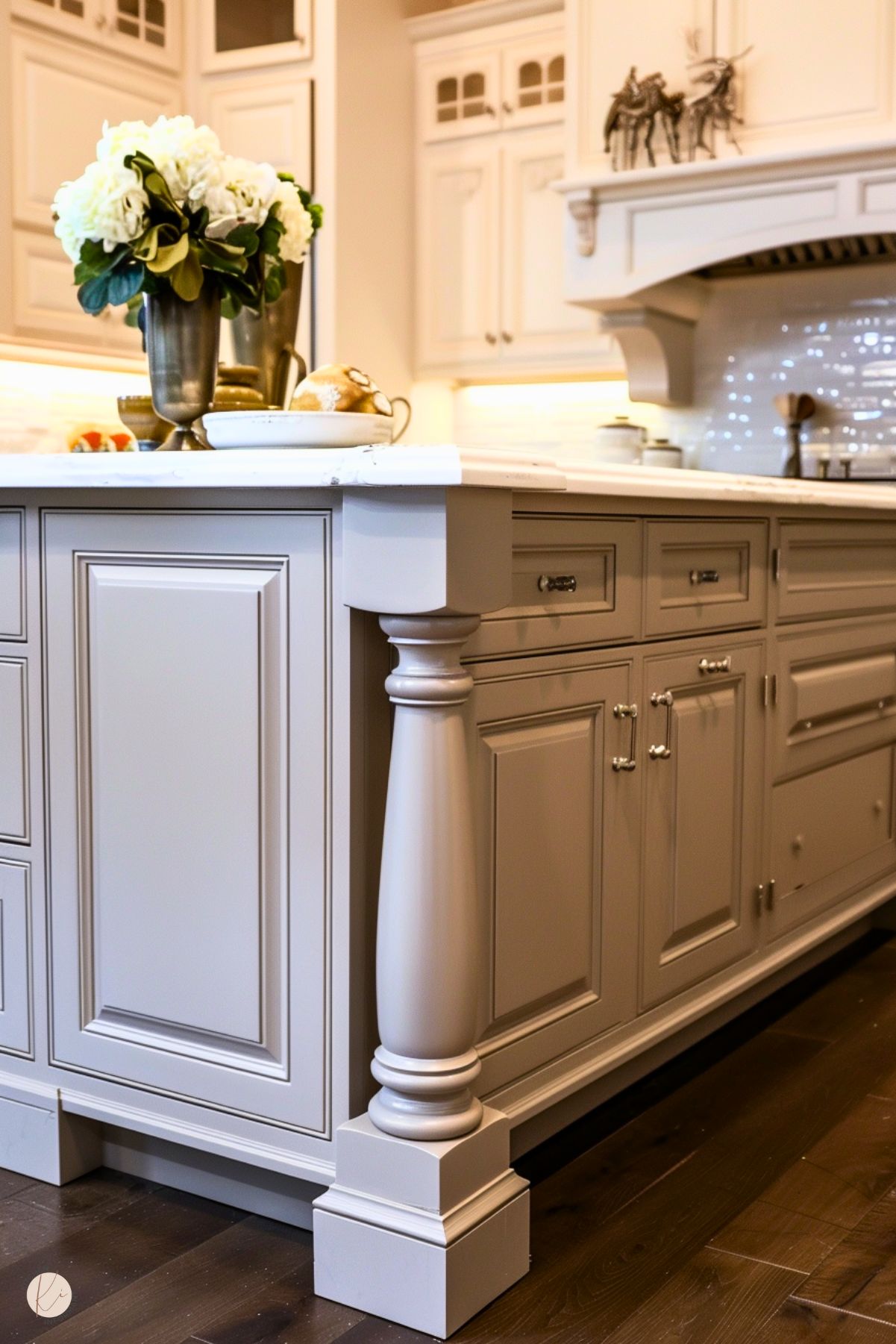 The image shows a greige kitchen island with a classic and elegant design, featuring detailed paneling and a decorative column on the corner. The countertop is white marble, styled with metallic vases holding white hydrangeas and a tray with decorative items. In the background, matching greige cabinetry and a white tiled backsplash are visible, complemented by warm under-cabinet lighting.