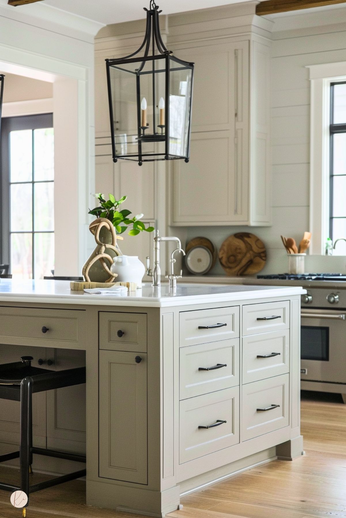 The image features a bright and airy kitchen with a greige island that has shaker-style cabinetry and black hardware. The island includes a white countertop, a polished silver faucet, and decorative items such as a white pitcher and greenery. A black metal lantern-style pendant light hangs above, adding a rustic charm. The background displays greige cabinetry, a stainless steel stove, and wooden accents like cutting boards, enhancing the warm, inviting atmosphere.