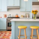 A bright kitchen with pastel blue cabinets, white countertops, and yellow barstools. Geometric-patterned floor tiles add a playful touch, while a warm-toned rug complements the space. Stainless steel appliances, an orange teapot, and fresh yellow flowers bring warmth and contrast.