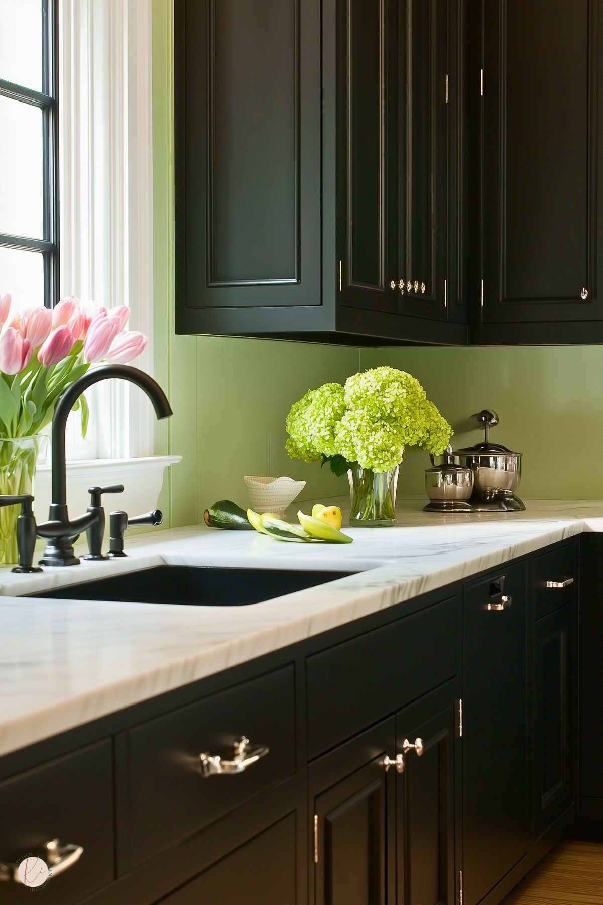 A charming kitchen with black cabinetry, white marble countertops, and a soft green backsplash that adds a refreshing touch. A black farmhouse-style faucet complements the dark sink. The countertop is adorned with a vase of pink tulips, a vase of green hydrangeas, and fresh produce, adding natural vibrancy to the space. Stainless steel appliances and silver hardware enhance the modern yet inviting design.