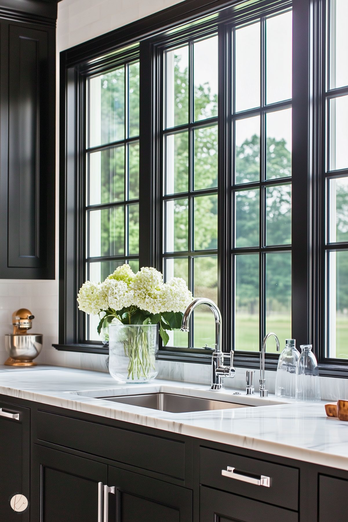 A bright kitchen featuring black cabinetry with a white marble countertop. The large window with black trim provides a view of lush greenery, flooding the space with natural light. A polished chrome faucet and a double sink sit beneath the window. A vase of white hydrangeas adds a touch of freshness and elegance, while a gold stand mixer in the corner subtly complements the clean, sophisticated design.