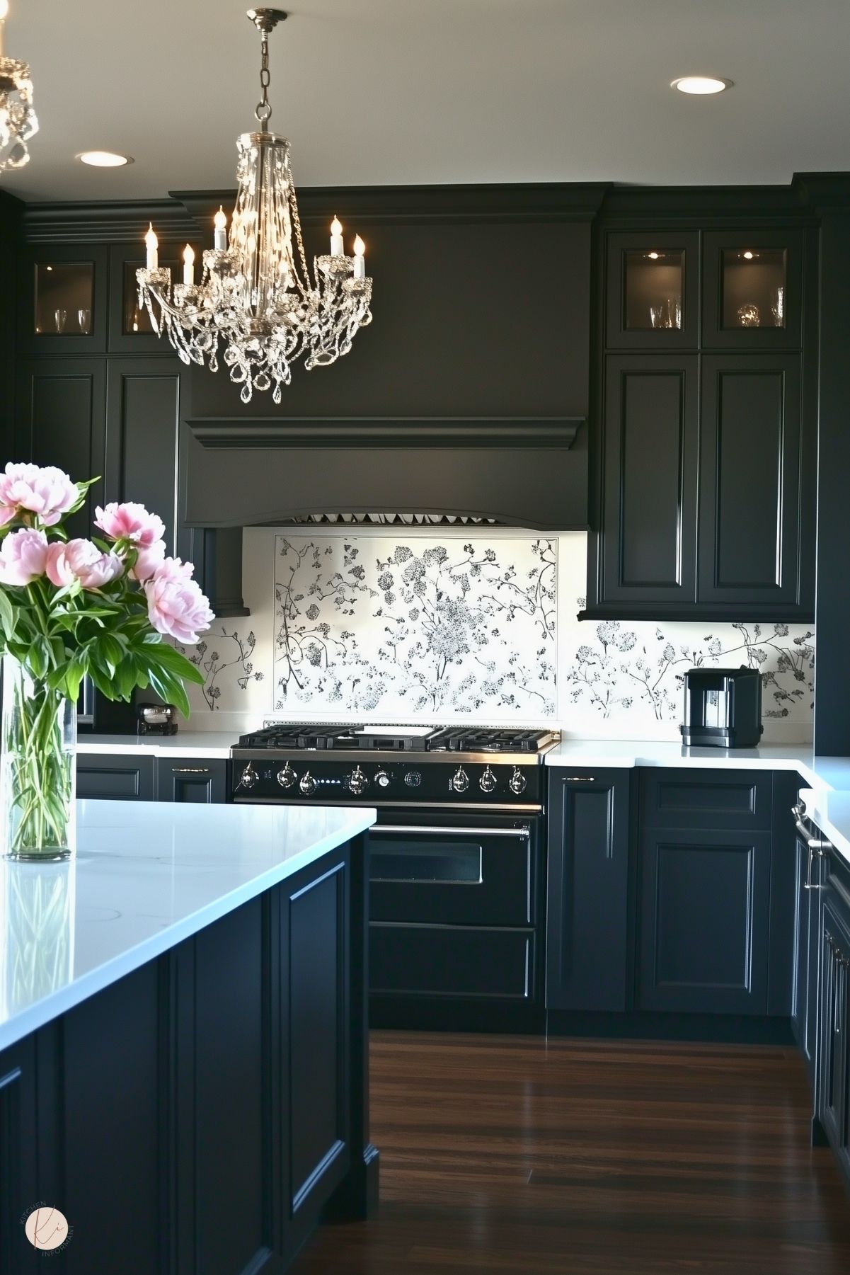 A sophisticated kitchen featuring black cabinetry, a matching black range with chrome details, and a white backsplash adorned with intricate black floral patterns. A crystal chandelier hangs elegantly above the space, adding a touch of luxury. White countertops provide a clean contrast, and a vase of pink peonies brings a soft, colorful accent to the dark and refined design. The polished wood flooring completes the classic aesthetic.