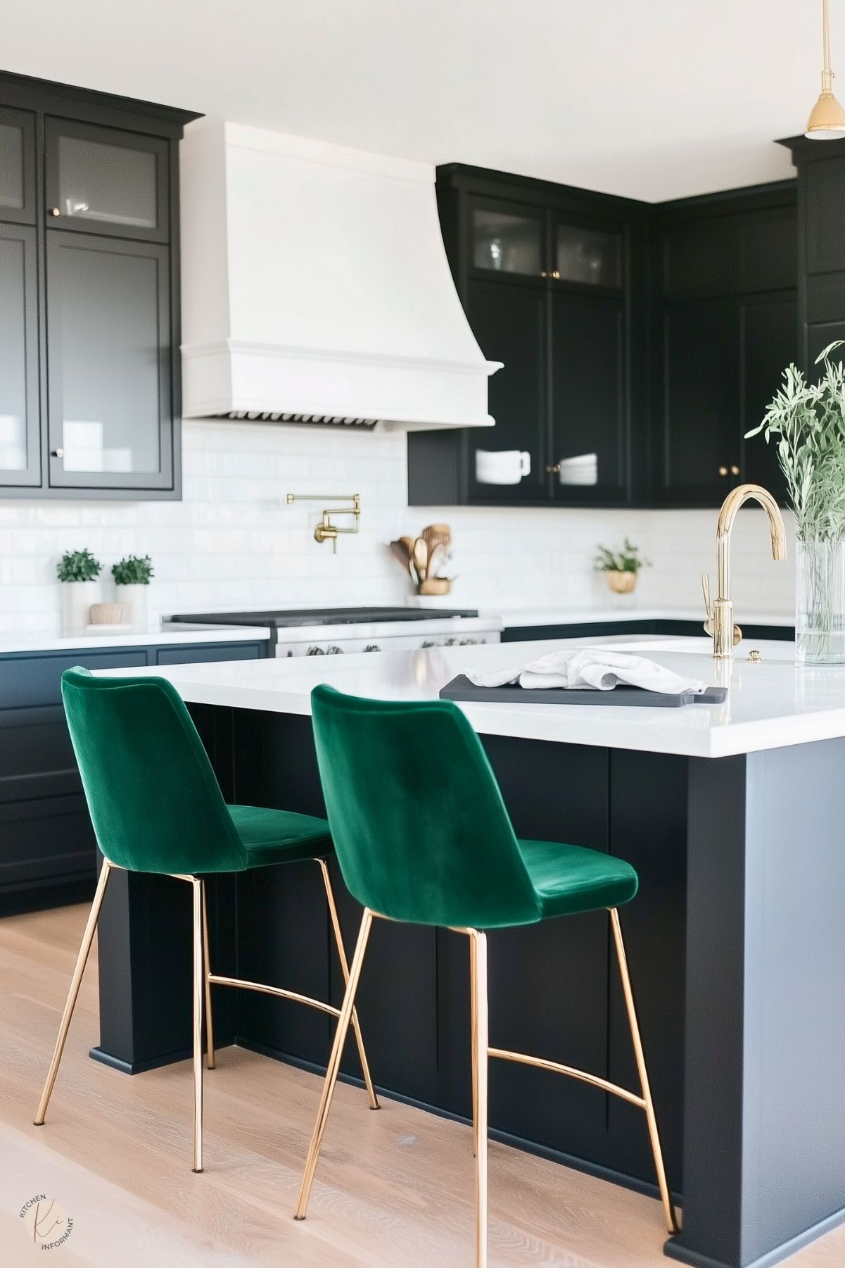 A modern kitchen with black cabinetry, a white marble countertop, and a bold white range hood. The space is accentuated by emerald green velvet chairs with gold legs, adding a touch of luxury and color. The backsplash features classic white subway tiles, complemented by brass fixtures, including a sleek faucet and pot filler. Light wood flooring and small potted plants bring warmth and freshness to the elegant design.