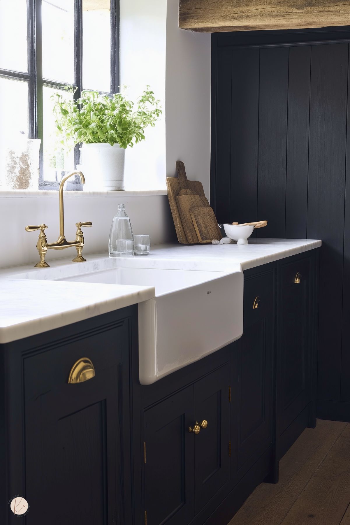 A cozy, rustic kitchen featuring black cabinetry with brass hardware and a white farmhouse sink. The countertop is a clean white marble, accented by a brass faucet that adds a touch of elegance. A potted basil plant sits on the windowsill, bringing a fresh, natural element to the space. Wooden cutting boards and a simple white bowl add warmth, while the black-paneled wall and exposed wooden beam enhance the farmhouse charm.