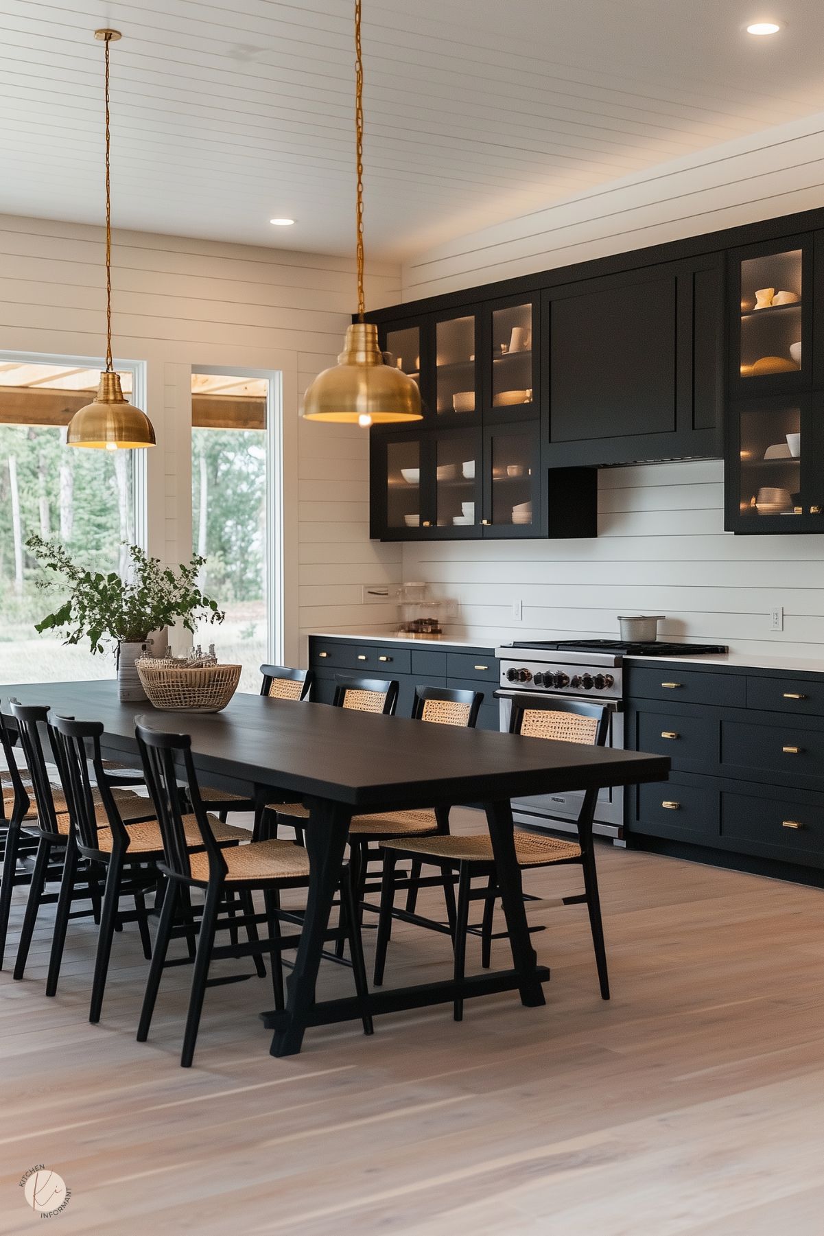 A farmhouse-style kitchen with black cabinetry, glass-front upper cabinets, and brass hardware. A large black dining table with matching chairs featuring woven seats anchors the space. The shiplap walls and light wood flooring create a warm and inviting atmosphere. Two brass pendant lights hang above the table, adding elegance, while large windows provide natural light and a view of the outdoors. The open shelving displays simple, minimalist dishware.