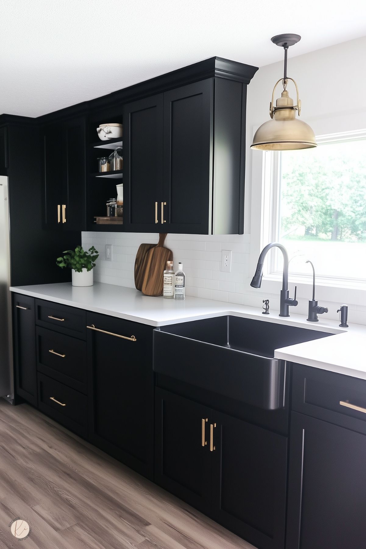 A modern farmhouse kitchen featuring black cabinetry with brass hardware and a deep black farmhouse sink. The countertops are clean white, paired with a classic white subway tile backsplash. A brass pendant light hangs above the sink, adding warmth and elegance. Open shelving displays jars and kitchen essentials, while a wooden cutting board and a potted plant add natural elements to the bright and airy space. The light wood flooring completes the look.