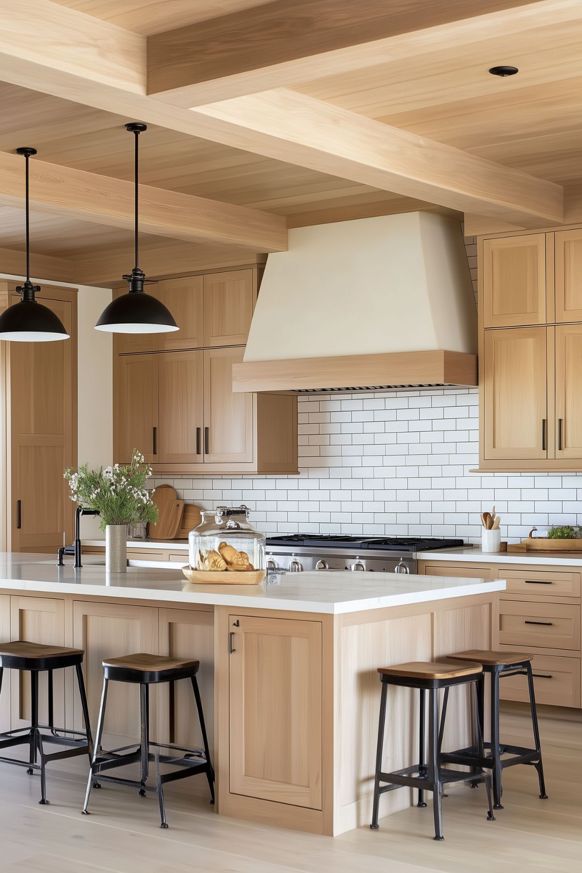 A bright modern farmhouse kitchen with light natural wood cabinetry and a large island topped with a white countertop. Black metal stools with wooden seats provide seating at the island, while black industrial pendant lights hang above. The backsplash features classic white subway tiles, and a simple wood-trimmed range hood complements the cabinetry. The space is styled with minimal decor, including fresh greenery and baked goods, creating a clean, inviting atmosphere.