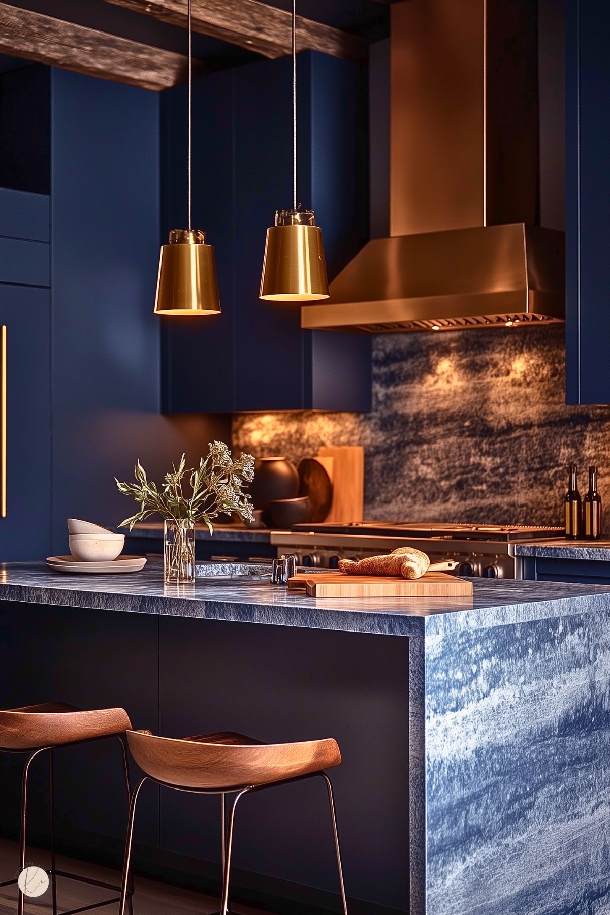 A striking kitchen with deep navy cabinetry and a bold marble countertop and backsplash in shades of blue and gray. The space features a brushed gold range hood and matching pendant lights that add warmth and elegance. Wooden barstools with curved seats provide a natural contrast, while minimal decor, including a vase with dried flowers and stacked ceramic bowls, creates a clean, sophisticated aesthetic. Exposed wooden ceiling beams enhance the cozy yet modern design.