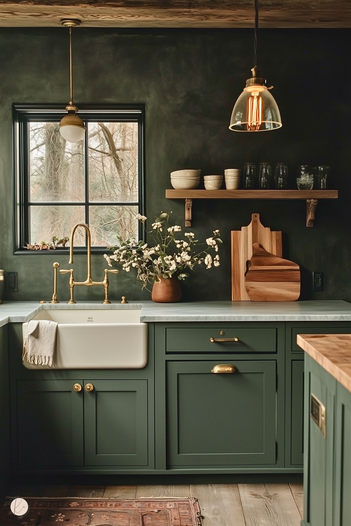 A charming kitchen with deep green cabinetry, brass hardware, and a farmhouse sink with a vintage-style brass faucet. The countertop is light marble, complemented by a wooden floating shelf displaying rustic bowls and glasses. A terra-cotta vase with white flowers adds a soft, natural touch. The walls have a textured finish, and a black-framed window brings in natural light. Pendant lighting with brass and glass accents enhances the warm and inviting ambiance. A patterned rug and wooden ceiling beams complete the cozy look.