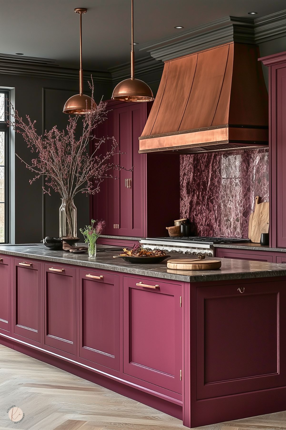 A bold kitchen design with deep burgundy cabinetry featuring brass handles and a dark gray stone countertop. The backsplash and range hood area are highlighted by matching burgundy marble and a striking copper range hood. Pendant lights with a copper finish hang above, adding warmth. A centerpiece vase with blooming branches enhances the luxurious, moody atmosphere, complemented by a chevron-patterned light wood floor.