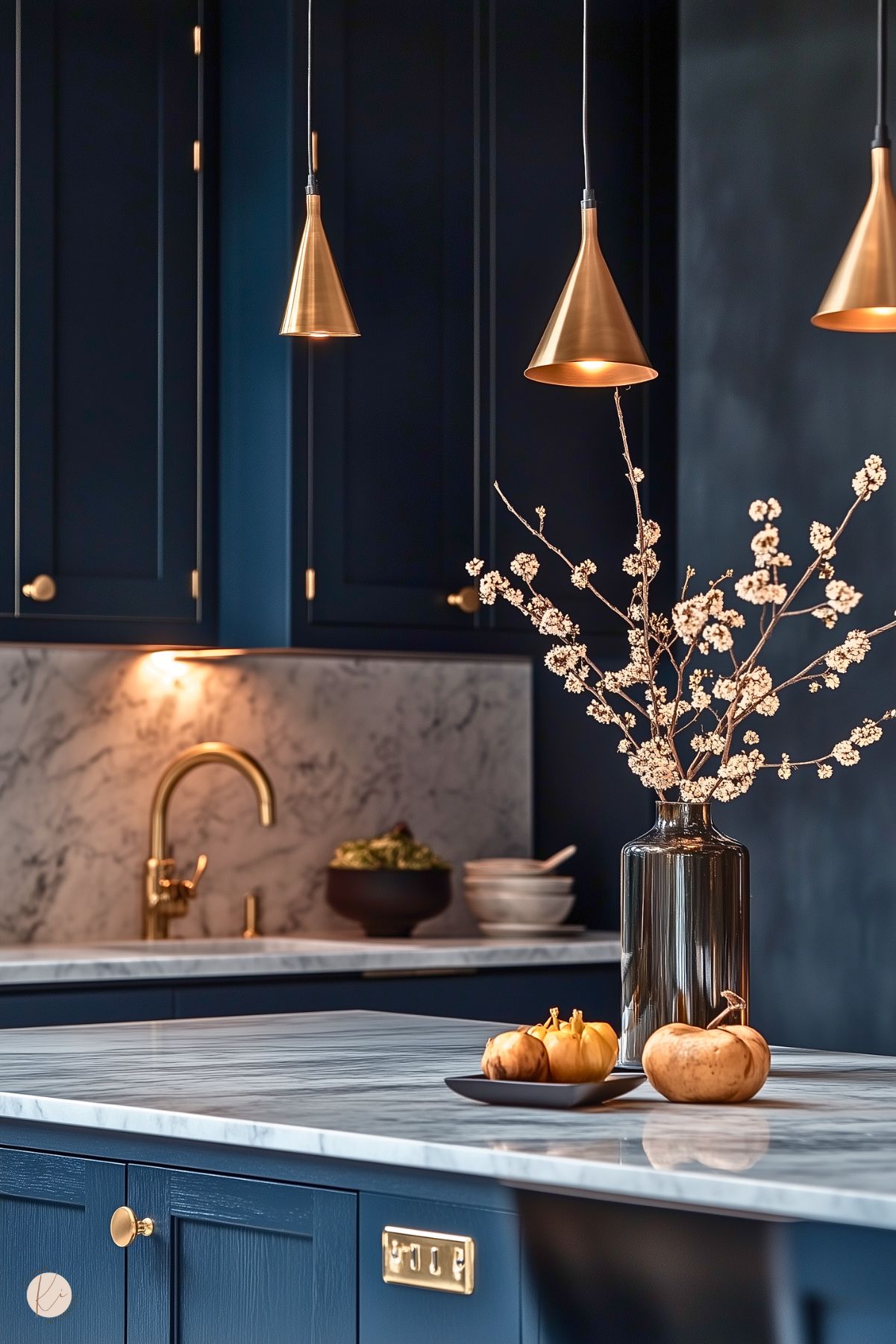 An elegant kitchen featuring deep navy cabinetry with gold hardware and white marble countertops and backsplash. Sleek cone-shaped pendant lights in brushed gold add a warm glow. The centerpiece is a marble-topped island adorned with a black vase holding delicate white blossoms and a small dish of decorative gourds. A gold faucet and minimalist decor elements enhance the sophisticated and luxurious design.