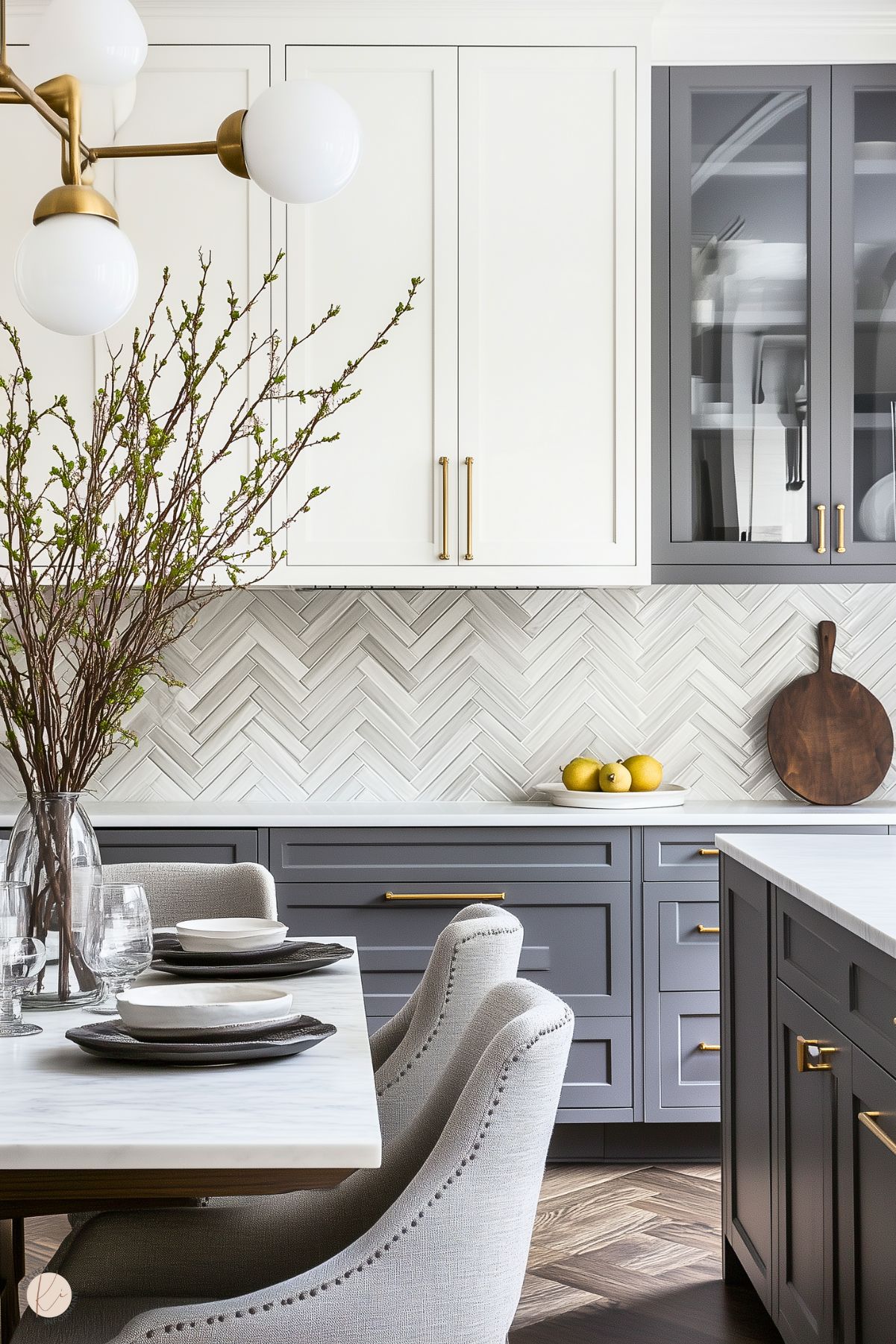 A sophisticated kitchen with a two-tone cabinetry design, featuring white upper cabinets and gray lower cabinets, both with brass hardware. The backsplash showcases a white herringbone tile pattern, adding texture and elegance. A dining area with upholstered chairs and a marble-topped table is set with modern dishware. Decorative branches in a glass vase and lemons on the counter provide natural accents, complementing the warm lighting and chevron-patterned wood flooring.