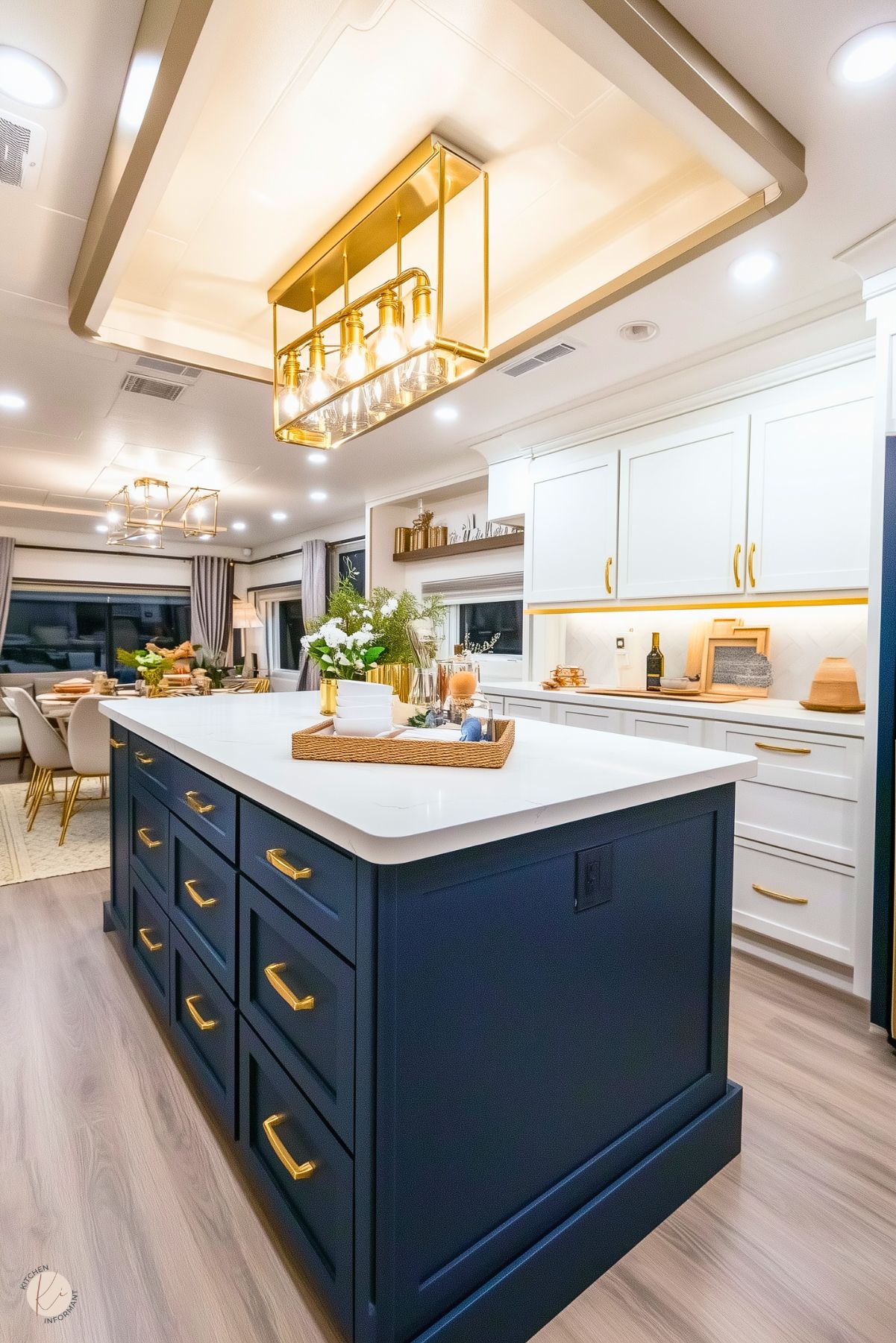 A stylish and modern kitchen featuring a navy blue island with gold hardware and a white quartz countertop. Overhead, a gold rectangular light fixture with exposed bulbs adds a contemporary touch. The surrounding cabinetry is white with gold accents, complemented by a well-lit backsplash. The open space extends into a dining area with elegant furnishings and decor, creating a cohesive and inviting atmosphere.








