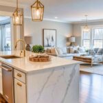 A bright, open-concept kitchen and living space featuring a white island with a waterfall marble countertop, brushed gold fixtures, and pendant lighting with geometric glass frames. The living area has a neutral color palette with a cozy sectional, large windows, and decor elements like a wooden coffee table and modern artwork on the wall. Warm wood flooring ties the spaces together.