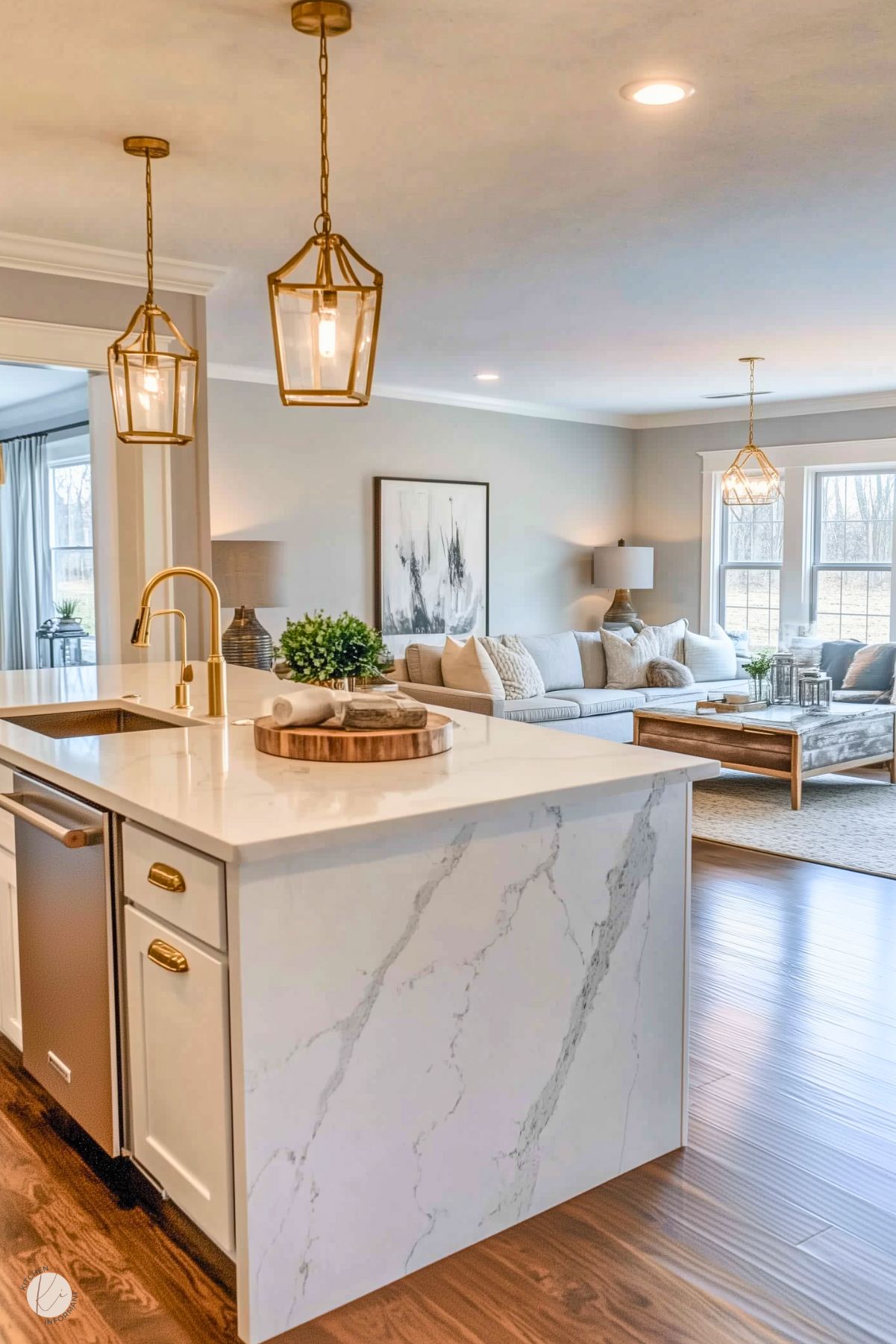 A bright, open-concept kitchen and living space featuring a white island with a waterfall marble countertop, brushed gold fixtures, and pendant lighting with geometric glass frames. The living area has a neutral color palette with a cozy sectional, large windows, and decor elements like a wooden coffee table and modern artwork on the wall. Warm wood flooring ties the spaces together.