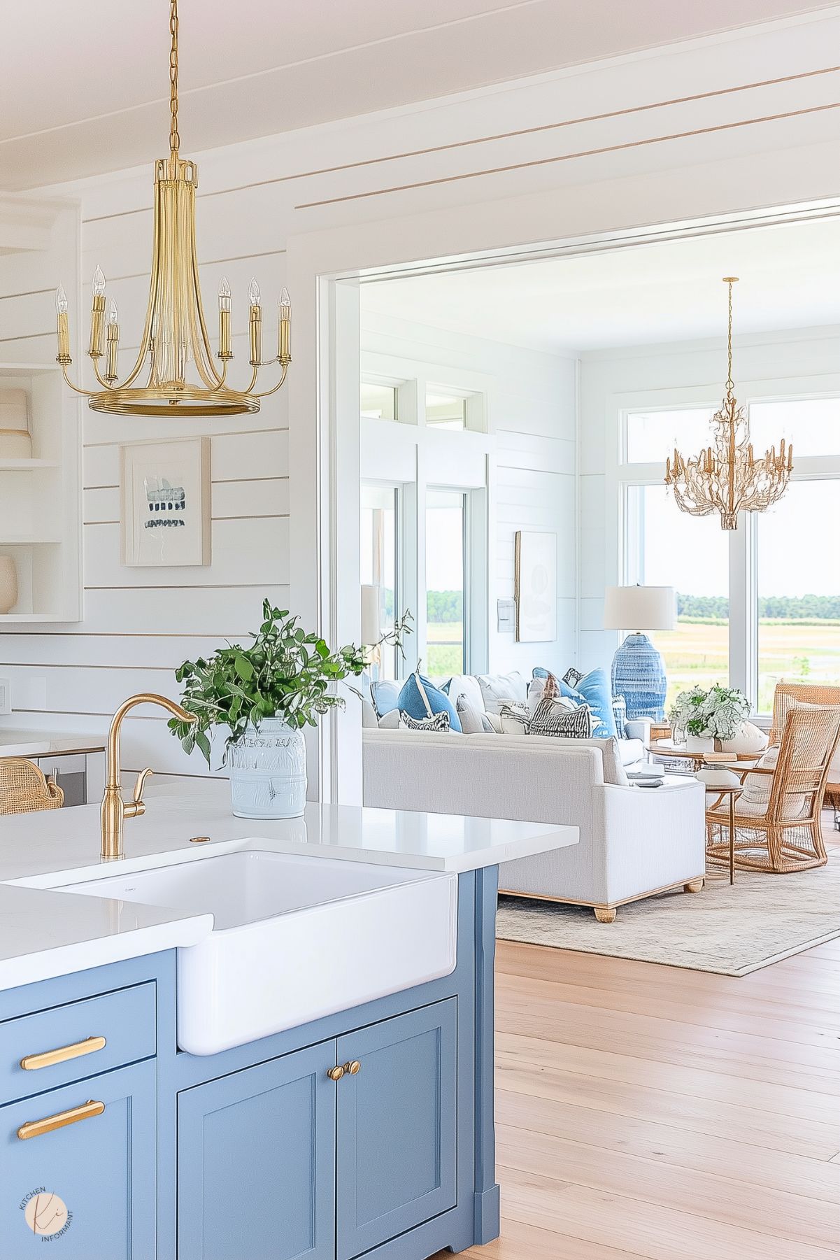 A coastal-inspired open-concept kitchen and living space with soft, natural tones. The kitchen features a farmhouse sink, a blue cabinetry island with gold hardware, and a brass faucet. Elegant chandeliers add sophistication, while white shiplap walls and large windows flood the area with natural light. The adjacent living room includes a white sofa, blue accents, wicker chairs, and a serene view of the outdoors.