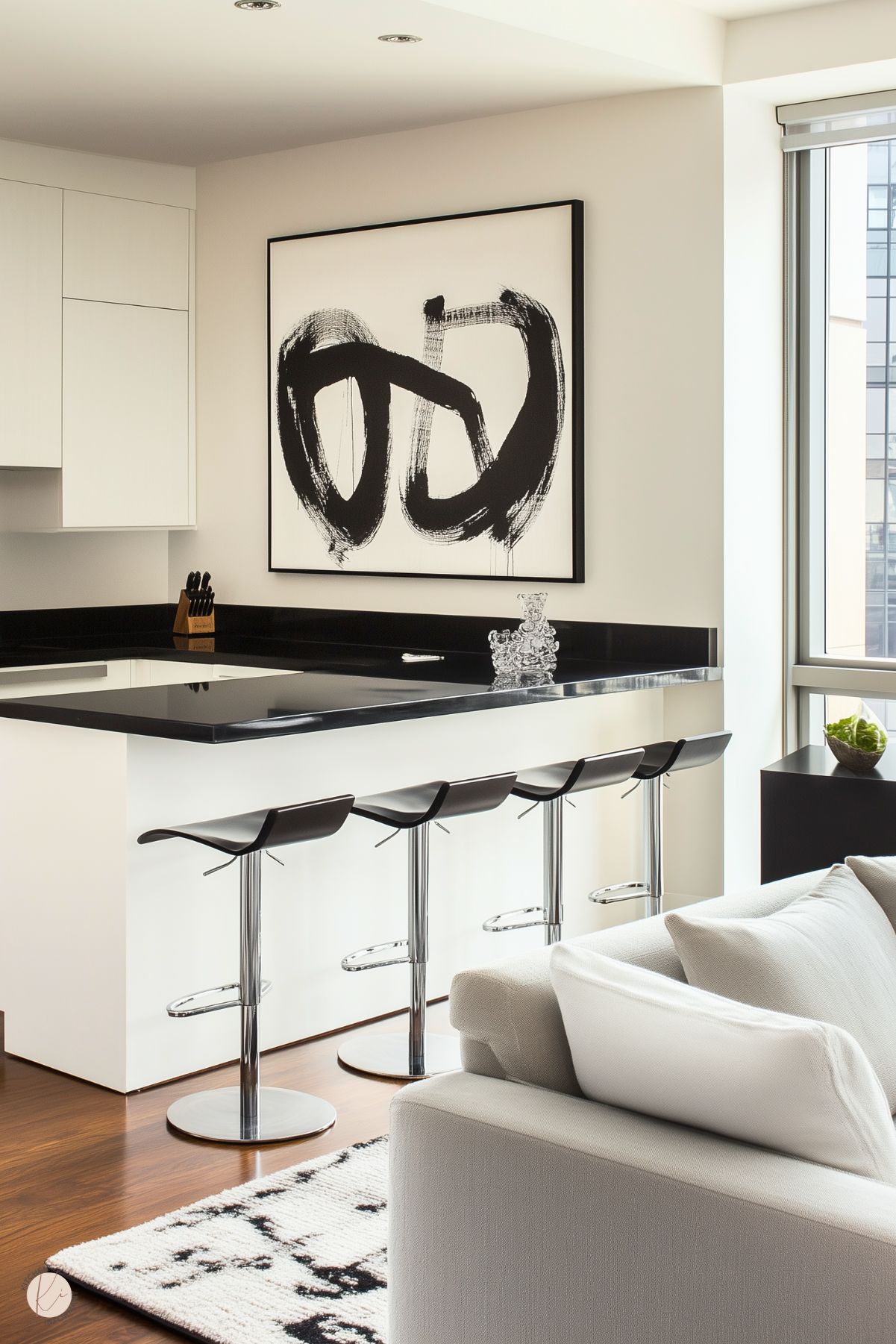 A modern kitchen and living area with a sleek black-and-white color scheme. The kitchen features a glossy black countertop with white cabinetry, complemented by minimalist bar stools with chrome bases and curved black seats. A bold abstract black-and-white artwork hangs on the wall, adding a contemporary touch. The living space includes a white sofa and a patterned rug, with warm wood flooring tying the design together.