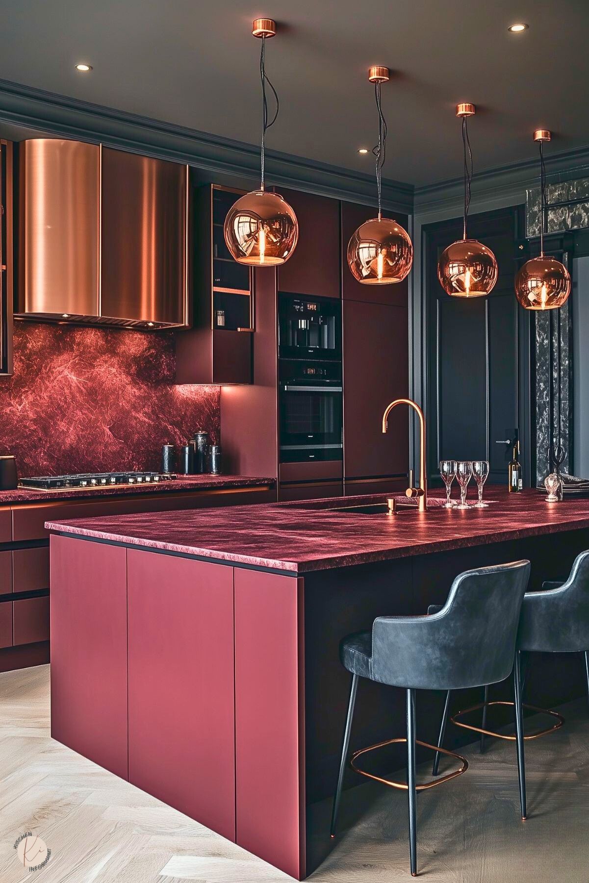 A luxurious modern kitchen with rich burgundy cabinetry and countertops accented by a matching marble backsplash. The centerpiece is a sleek island with a bold burgundy surface and integrated copper faucet, paired with black leather barstools featuring copper footrests. A row of polished copper pendant lights hangs above, adding warmth and elegance. The metallic elements and herringbone wood flooring enhance the sophisticated, contemporary design.