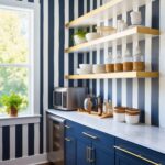 A playful preppy kitchen nook featuring navy and white striped walls, navy cabinetry with gold hardware, and open gold shelving. The space is accented with marble countertops, glass jars, and fresh greenery for a clean and vibrant look.